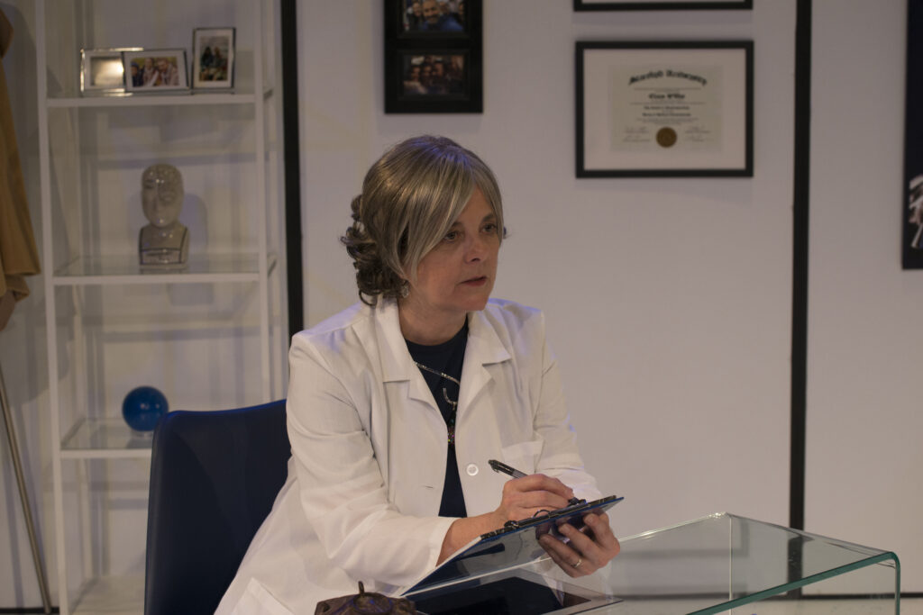 Still from 'Uncanny Valley'. Claire is sitting at a desk with a clipboard, taking notes about Julian.