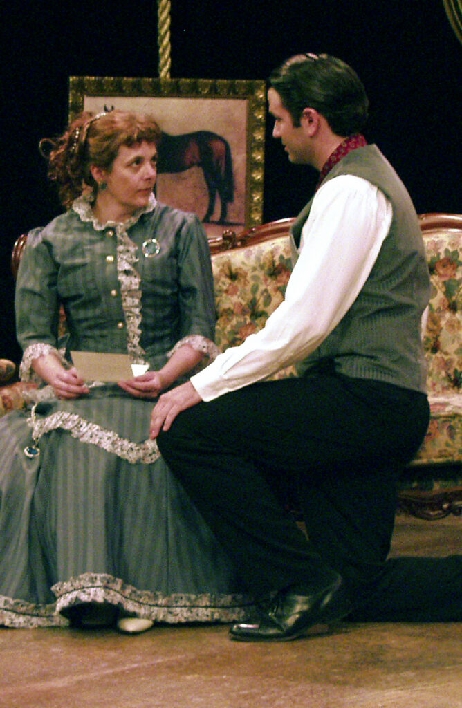 A scene from The Day After The Fair.A woman is sitting on a sofa and a man is kneeling before her. Both are dressed in Victorian clothing.