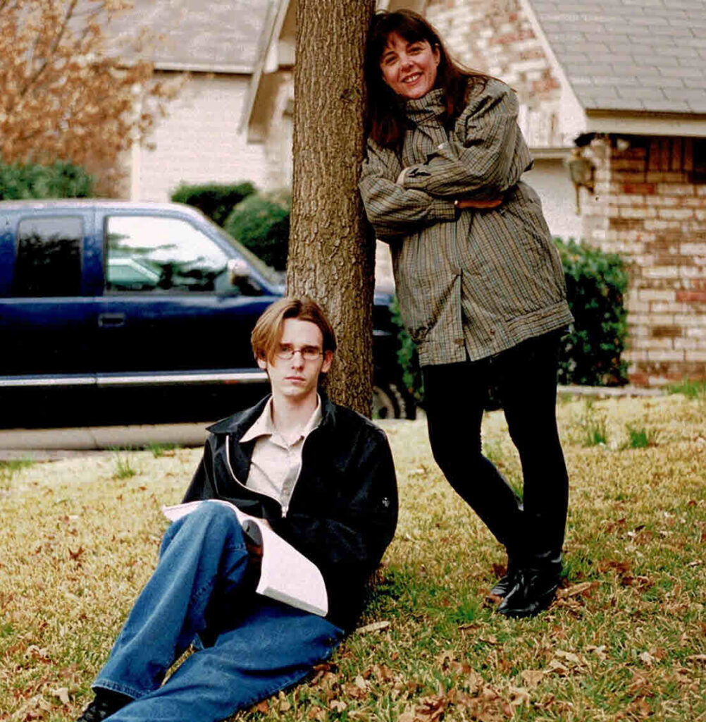 Sue standing leaning against a tree. Young man sitting on the ground. leaning up a against the tree, holding a script.