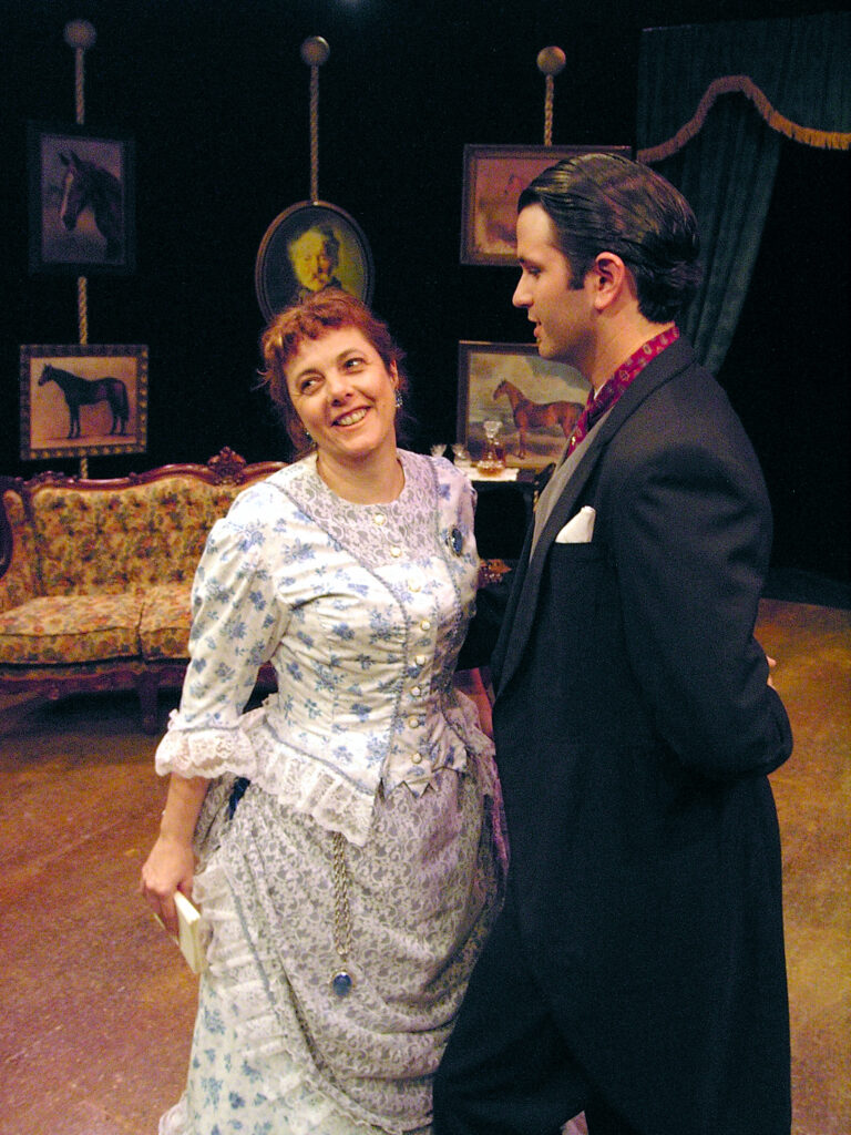 Woman and man in Victorian dress, standing on a set. Woman is laughing.