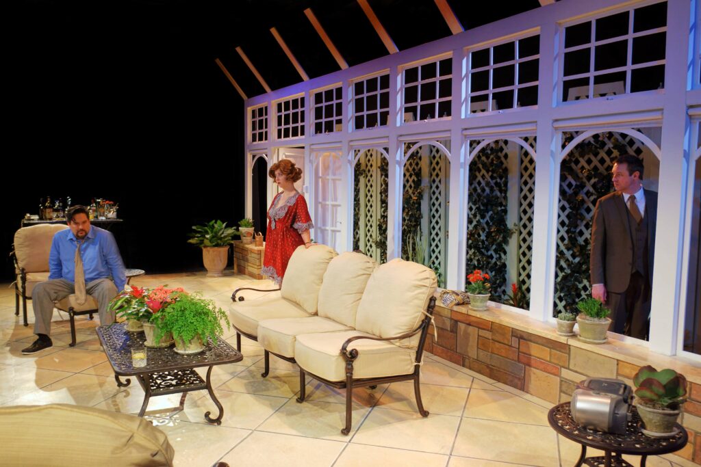 In a conservatory. Man sitting on chair with woman looking on. A man looking through the window at them.