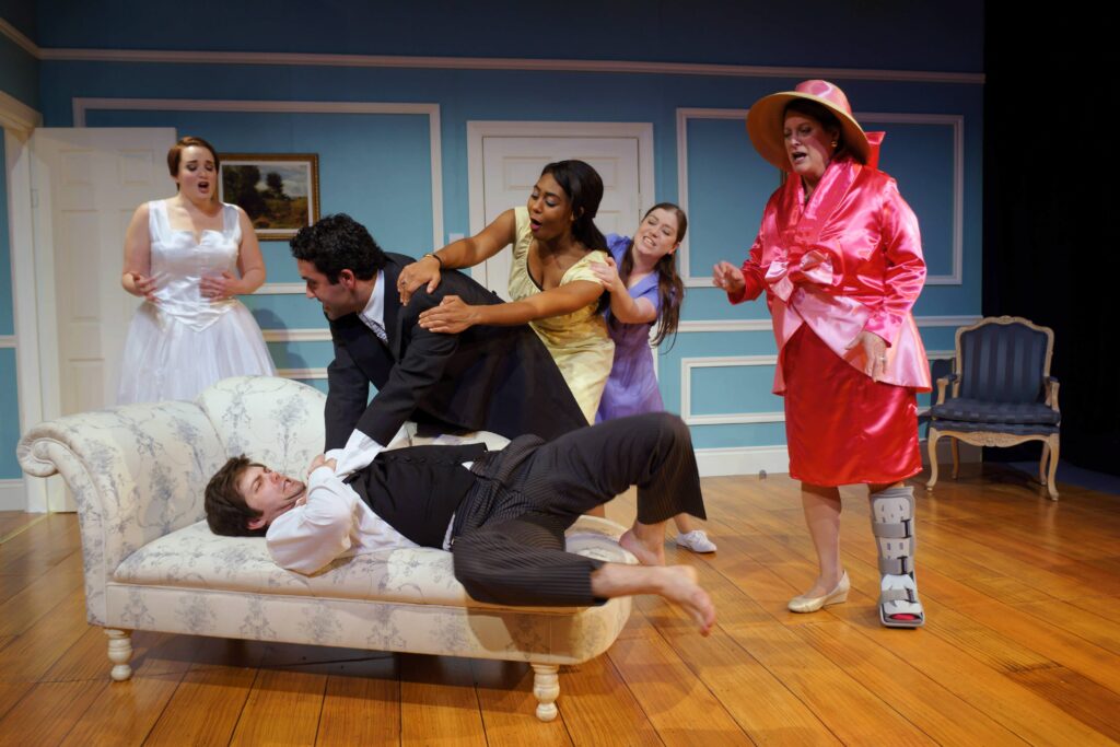 Man trying to strangle another man on a chaise longue, watched by four women dressed for a wedding.